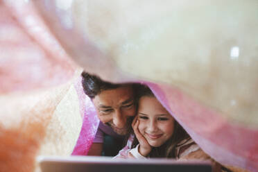 Happy daughter and father using tablet PC lying under blanket - JOSEF10376