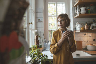 Mature woman with bowl standing in kitchen at home - JOSEF10377