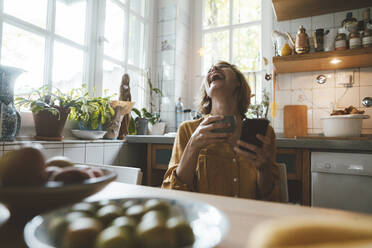 Fröhliche Frau mit Kaffeetasse und Smartphone am Esstisch sitzend - JOSEF10380