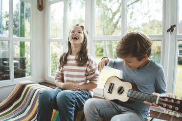 Cheerful girl sitting with brother playing guitar at home - JOSEF10395