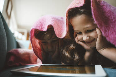 Glückliche Geschwister mit dem Kopf in den Händen, die einen Tablet-PC benutzen und zu Hause unter einer Decke liegen - JOSEF10396