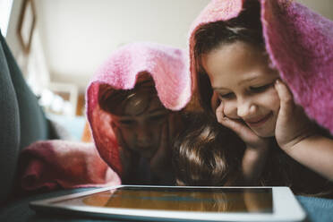 Happy siblings with head in hands using tablet PC lying under blanket at home - JOSEF10396