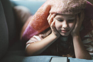 Lächelndes Mädchen mit Kopf in den Händen, das ein Smartphone benutzt und zu Hause unter einer Decke liegt - JOSEF10400