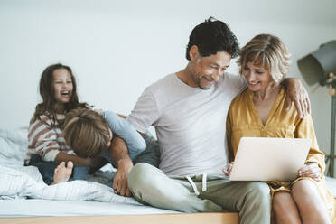 Happy man and woman with laptop sitting by daughter and son playing on bed at home - JOSEF10402