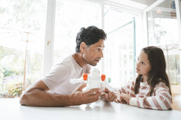 Daughter with father holding toy robot sitting at table - JOSEF10420