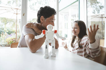 Happy daughter and father sitting with toy robot on table at home - JOSEF10422