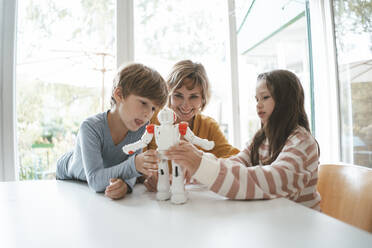 Girl and boy playing with toy robot sitting by mother at table - JOSEF10425