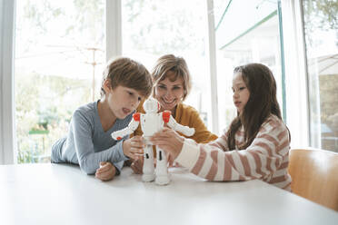 Mädchen und Junge spielen mit Spielzeugroboter bei der Mutter am Tisch sitzend - JOSEF10425