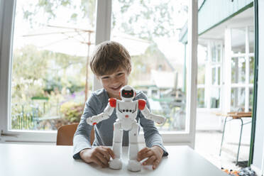 Glücklicher Junge mit Spielzeugroboter am Tisch sitzend - JOSEF10430