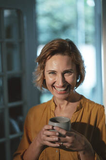 Glückliche reife Frau mit blondem Haar, die eine Kaffeetasse hält und zu Hause steht - JOSEF10439