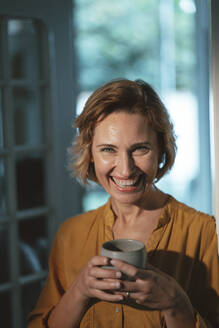 Happy mature woman with blond hair holding coffee cup standing at home - JOSEF10439