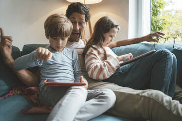 Happy father sitting with daughter and son using tablet PC at home - JOSEF10452