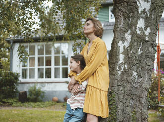 Smiling daughter with mother standing by tree trunk in back yard - JOSEF10492