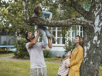 Frau und Mädchen sehen einen Mann, der einem Jungen beim Klettern auf einen Baum im Hinterhof hilft - JOSEF10493