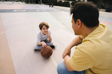 Junge mit Basketball vor seinem Vater auf dem Sportplatz sitzend - MMPF00107