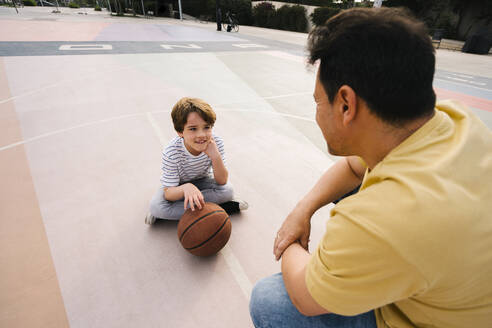 Boy with basketball sitting in front of father at sports court - MMPF00107