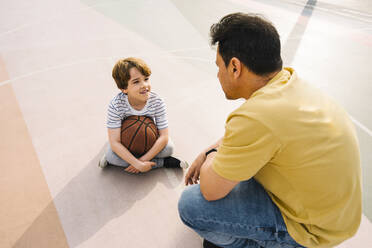 Boy with basketball sitting in front of father at sports court on sunny day - MMPF00108