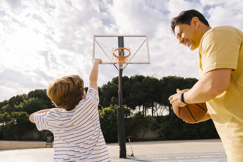 Playful boy with father holding basketball at sports court on sunny day - MMPF00110
