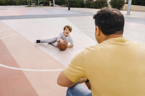 Happy boy with basketball sitting in front of father at sports court - MMPF00115