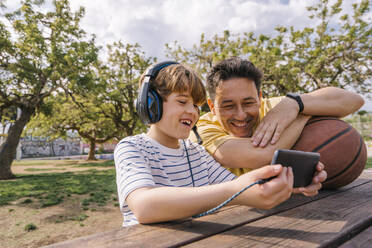 Smiling boy listening music through headphones sharing mobile phone with father sitting at table - MMPF00118