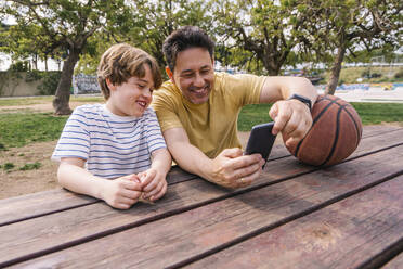 Lächelnder Mann mit Basketball, der seinem am Tisch sitzenden Sohn das Mobiltelefon zeigt - MMPF00119