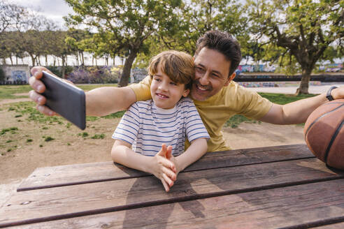 Smiling man with son taking selfie through smart phone sitting at table - MMPF00120