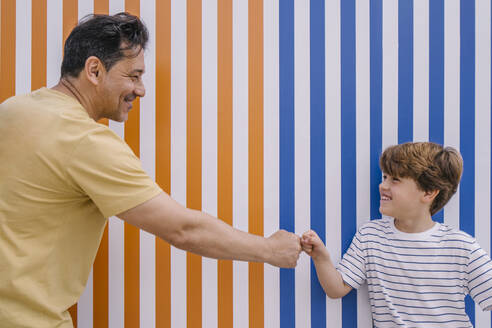 Smiling man giving fist bump to son in front of orange and blue striped wall - MMPF00122