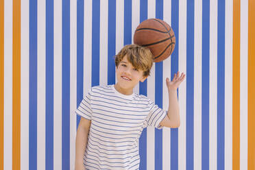 Playful boy balancing basketball on head in front of blue and orange striped wall - MMPF00124