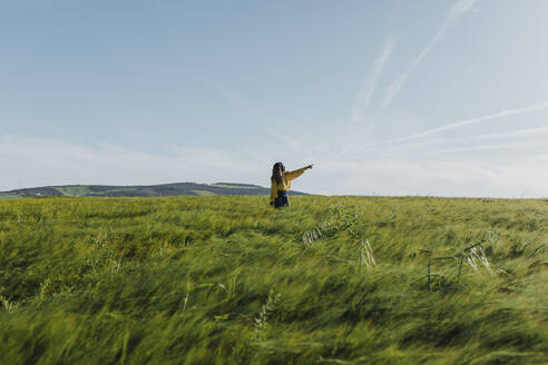 Junge Frau steht gestikulierend auf einer Wiese an einem sonnigen Tag - DMGF00737