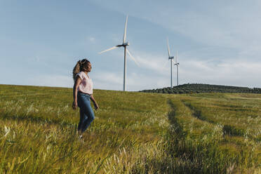 Lächelnde junge Frau, die auf einer Wiese vor einer Windkraftanlage spazieren geht - DMGF00754
