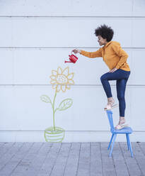 Woman standing on chair watering flowers painted on wall - UUF26431