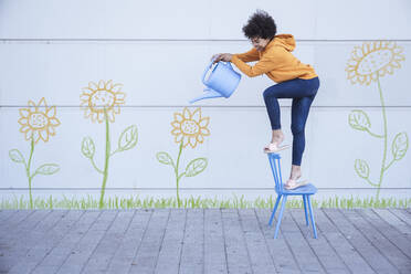 Woman standing on chair watering flowers painted on wall - UUF26434