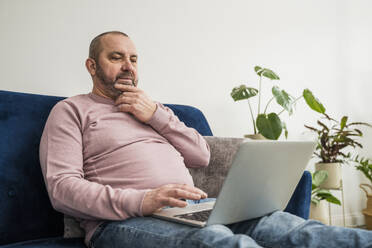 Nachdenklicher Geschäftsmann mit Laptop auf dem Sofa im Heimbüro sitzend - OSF00022