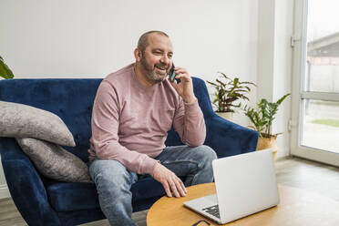Lächelnder Geschäftsmann, der mit seinem Laptop im Wohnzimmer sitzt und über sein Smartphone spricht - OSF00023
