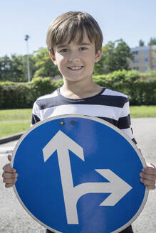 Smiling boy holding road sign board with arrow symbol on sunny day - RNF01394