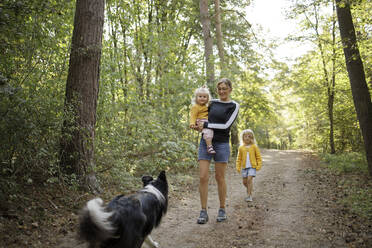 Mother carrying daughter walking together with girl and dog in forest - DWF00576