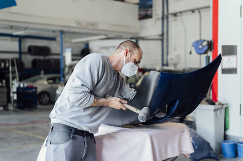 Mechaniker mit Schutzmaske hämmert in der Werkstatt an einer Karosserie - PGF01081