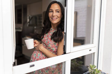 Smiling pregnant woman with tea mug standing by window - JCCMF06475