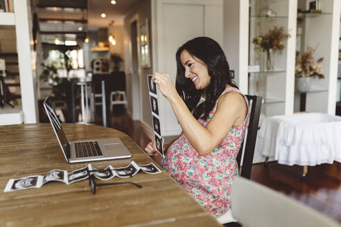 Glückliche schwangere Frau zeigt Ultraschallbilder per Videoanruf auf dem Laptop zu Hause - JCCMF06480