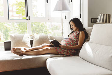 Lächelnde schwangere Frau, die auf dem Sofa im Wohnzimmer zu Hause ein Buch liest - JCCMF06481