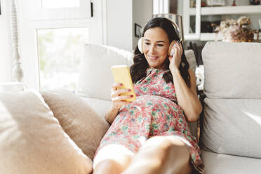 Lächelnde schwangere Frau, die über drahtlose Kopfhörer mit einem Smart-Pone auf dem Sofa zu Hause Musik hört - JCCMF06484