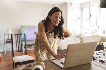 Smiling pregnant woman using laptop working from home - JCCMF06485