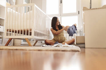 Lächelnde schwangere Frau mit Babykleidung an der Krippe zu Hause - JCCMF06487