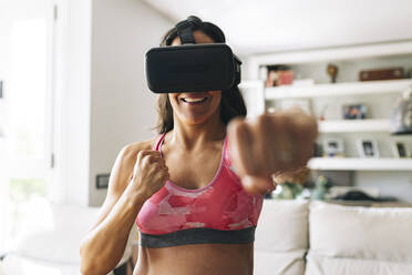 Smiling pregnant woman wearing virtual reality simulator punching at home - JCCMF06508