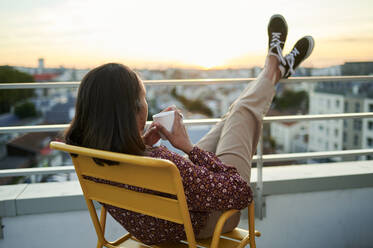 Junge Frau mit Kaffeetasse entspannt sich auf dem Balkon bei Sonnenuntergang - KIJF04481