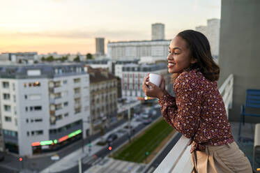 Lächelnde junge Frau mit Kaffeetasse auf dem Balkon bei Sonnenuntergang - KIJF04484