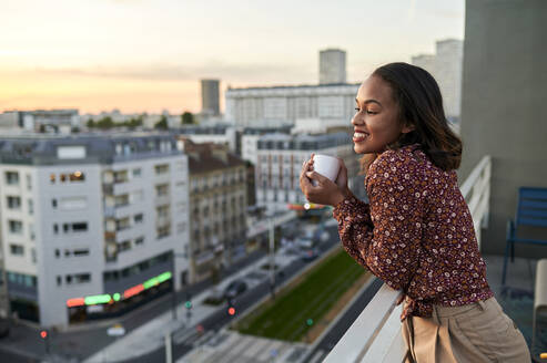 Lächelnde junge Frau mit Kaffeetasse auf dem Balkon bei Sonnenuntergang - KIJF04484