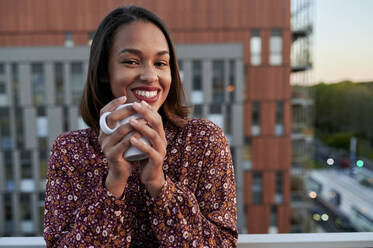 Happy young woman with coffee cup in front of building - KIJF04489
