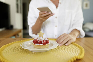 Woman photographing raspberry tart with star shape candle through smart phone at home - TYF00210