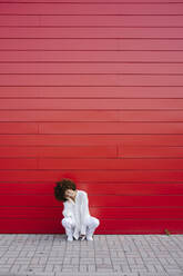 Young woman with head in hand crouching on footpath in front of red wall - GIOF15500